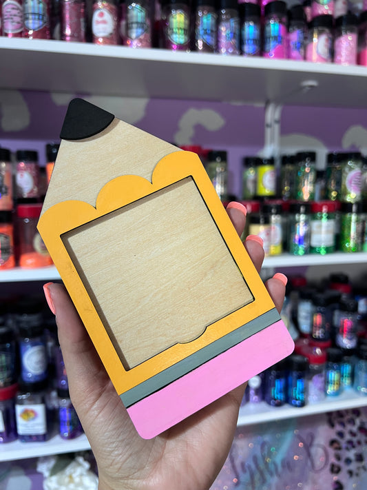 A wooden pencil-shaped post it note holder with a slot in the center, held in a hand, with shelves of various colorful items in the background.